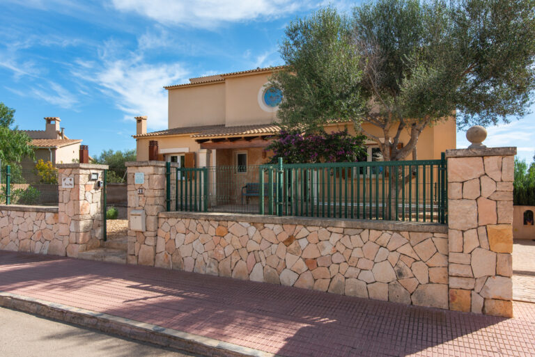 Straßenansicht der Villa Dues Illes in Portocolom mit Natursteinmauer, grünem Metallzaun, Toranlage und mediterraner Hausfassade