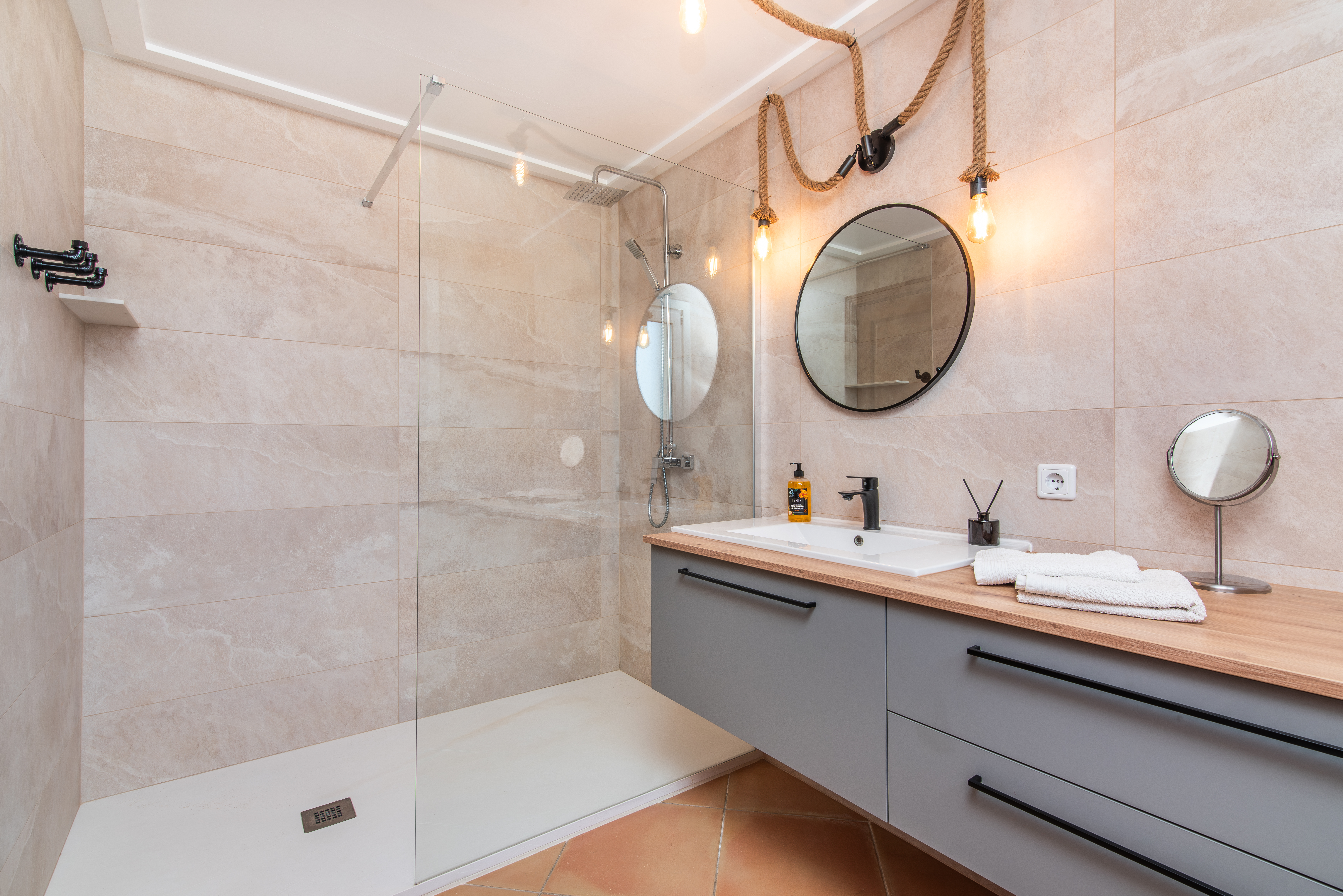 Badezimmer mit beigefarbenen Fliesen, großer Dusche, rundem Spiegel, hellgrauem Badmöbel mit weißem Waschbecken und Seilbeleuchtung