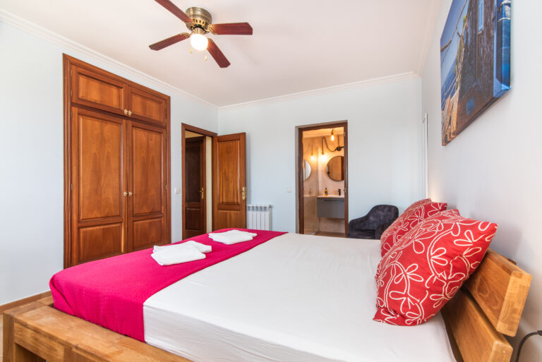 Helles Schlafzimmer mit massivem Holzbett, roter Tagesdecke, Deckenventilator, Einbauschrank und Blick ins angrenzende Badezimmer